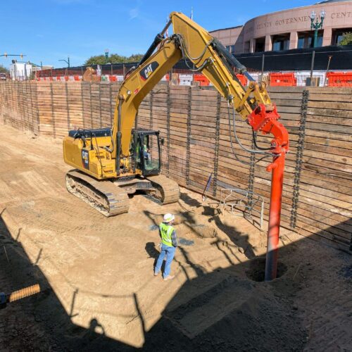 Worker guides yellow excavator with auger at site by walls.