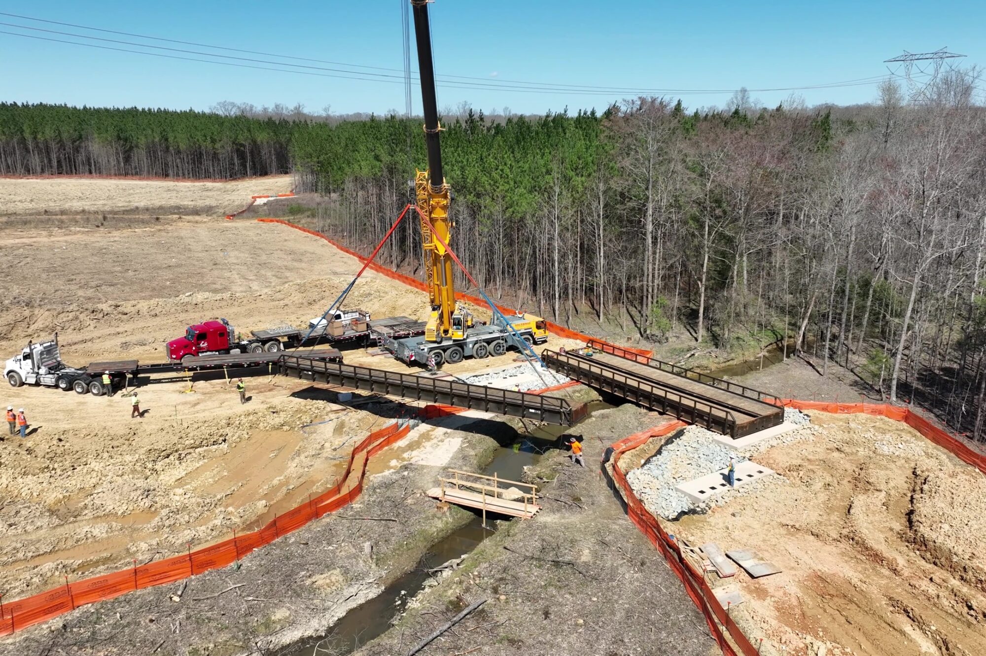 Crane lifts bridge section over stream at forested site.