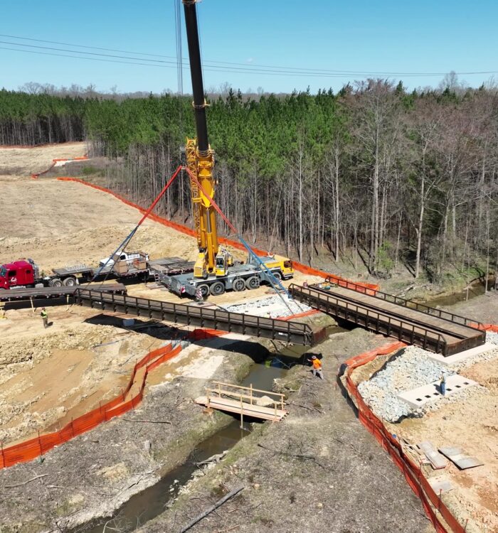 Crane lifts bridge section over stream at forested site.
