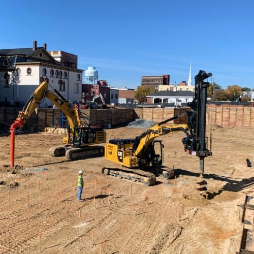Construction vehicles and worker on site with buildings, blue sky.