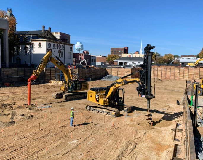 Construction vehicles and worker on site with buildings, blue sky.