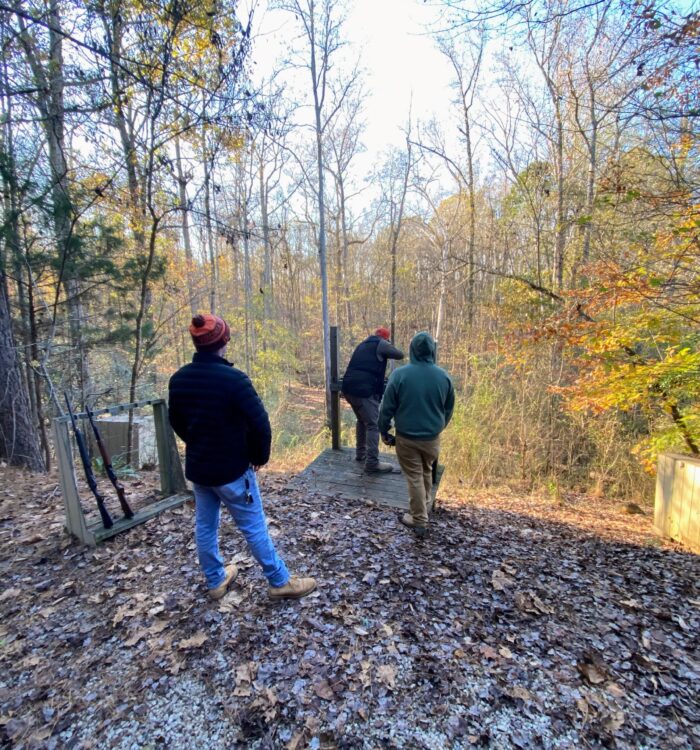 Three people on a wooden platform in a forest, one with shotgun.