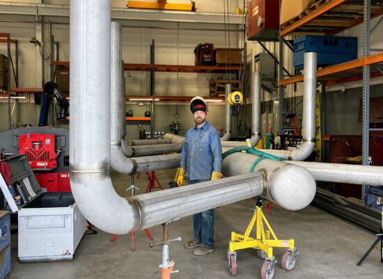 Man in safety gear in workshop with metal pipes and crane.