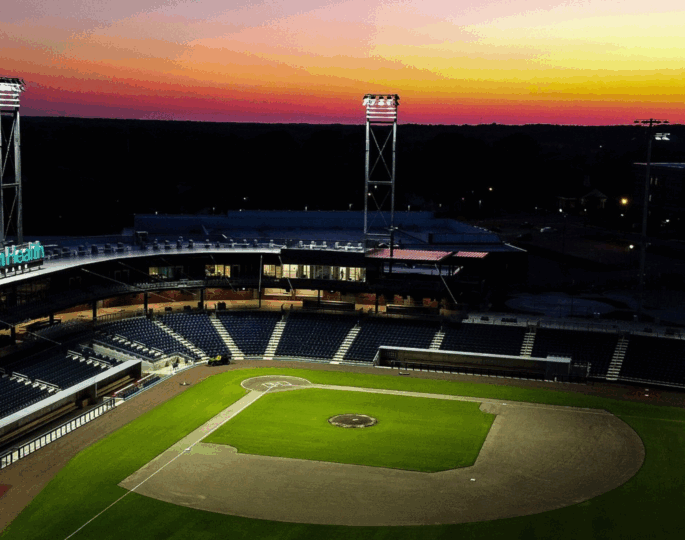 Empty baseball stadium at dusk, lights on, vibrant sky.