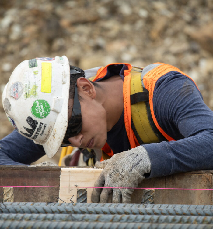 Worker in hard hat aligns wooden form; rebar and safety line visible.