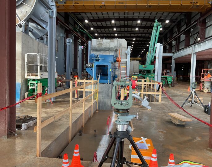 Indoor construction site with surveying gear, cones, railings, machinery.