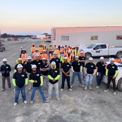 Construction workers in black shirts and orange vests pose outdoors.