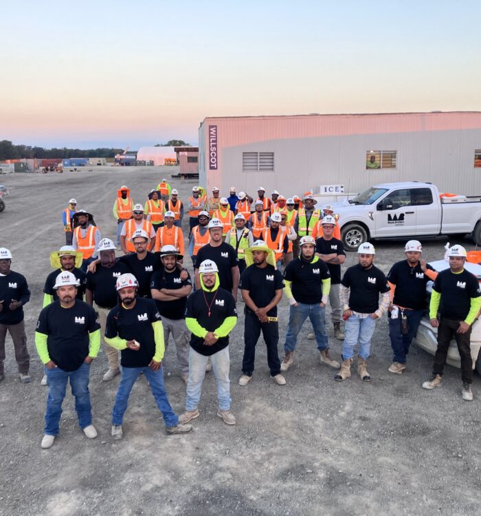 Construction workers in black shirts and orange vests pose outdoors.