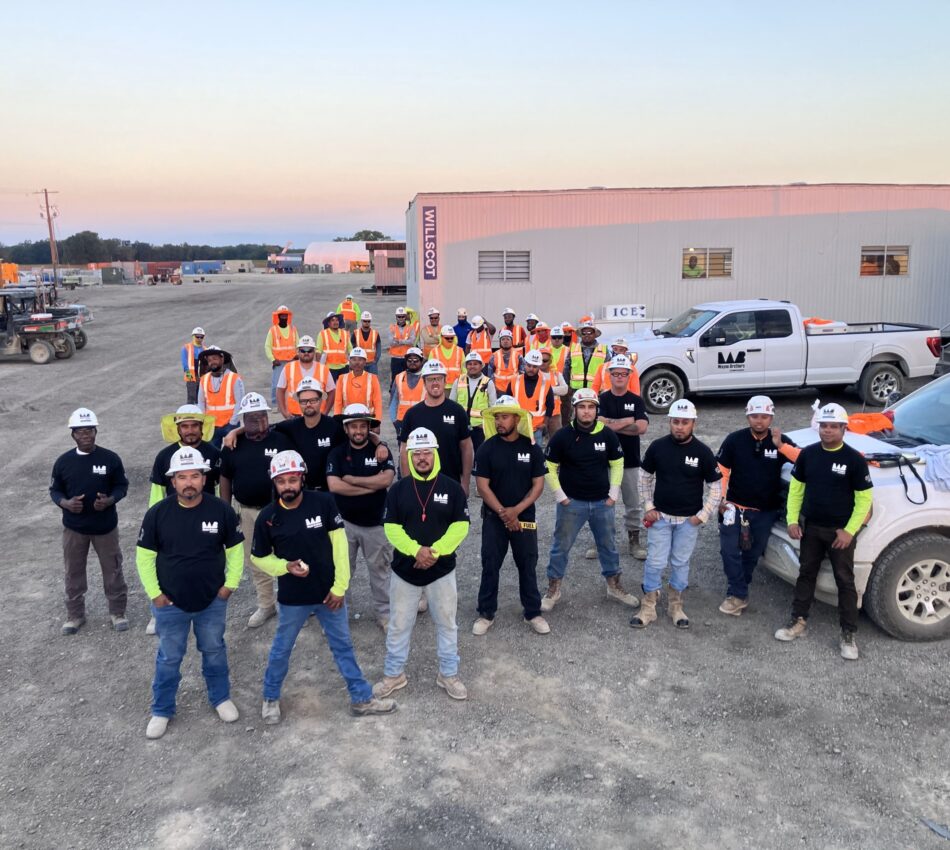 Construction workers in black shirts and orange vests pose outdoors.
