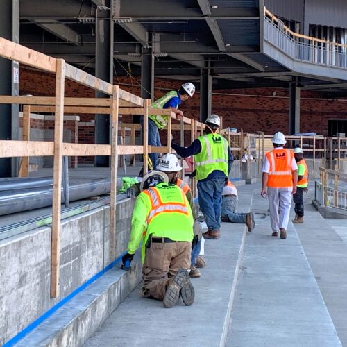 Workers in safety gear inspect and install structures by a walkway.