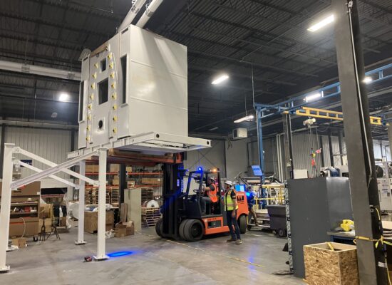 Workers use a forklift to move a large machine in a warehouse.