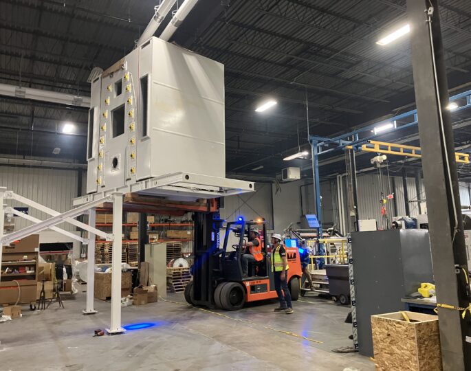 Workers use a forklift to move a large machine in a warehouse.