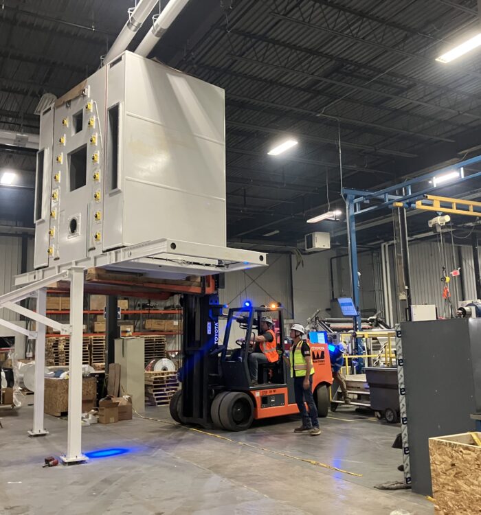 Workers use a forklift to move a large machine in a warehouse.