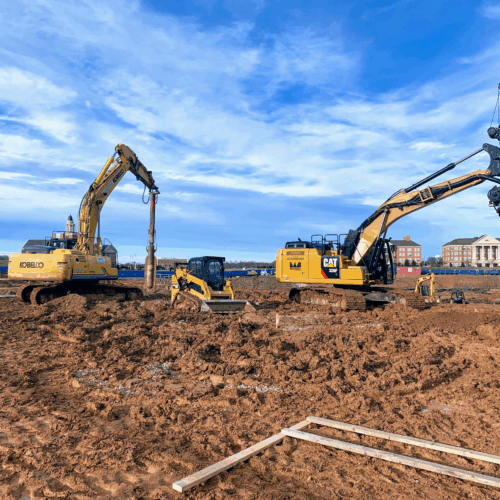 Excavators and bulldozer on muddy site with buildings behind.