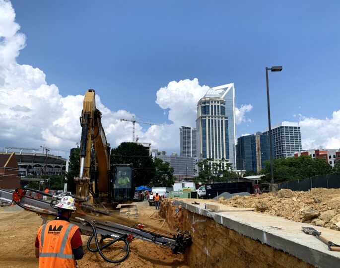 Workers operate machinery at city excavation site, skyscrapers behind.