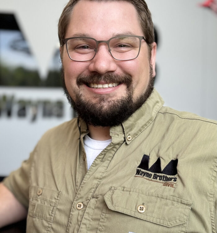 Bearded man with glasses smiles in tan Wayne Brothers shirt.