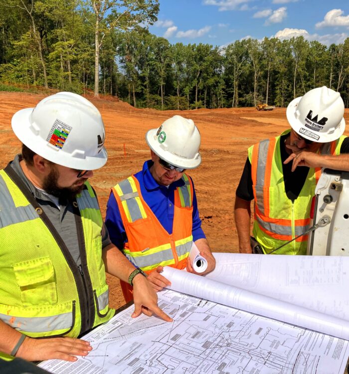 Three workers in vests and hard hats review a blueprint outside.
