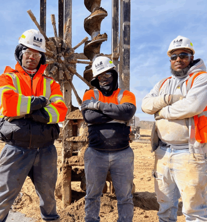 Three workers in safety gear stand by a large drill.