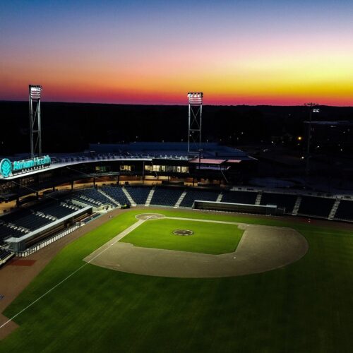 Empty baseball stadium at sunset with lights and glowing sign.