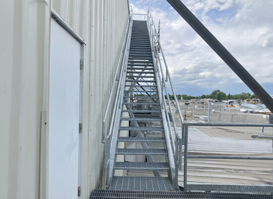 Metal staircase on industrial building with rooftops and cloudy sky.