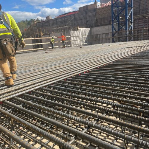 Workers stand on steel rebar grid at construction site, crane behind.