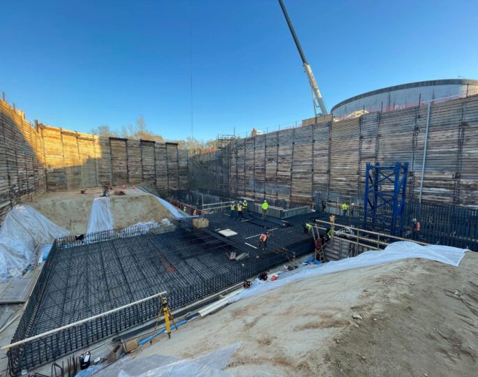 Workers assemble rebar in deep pit, wooden walls, clear sky.